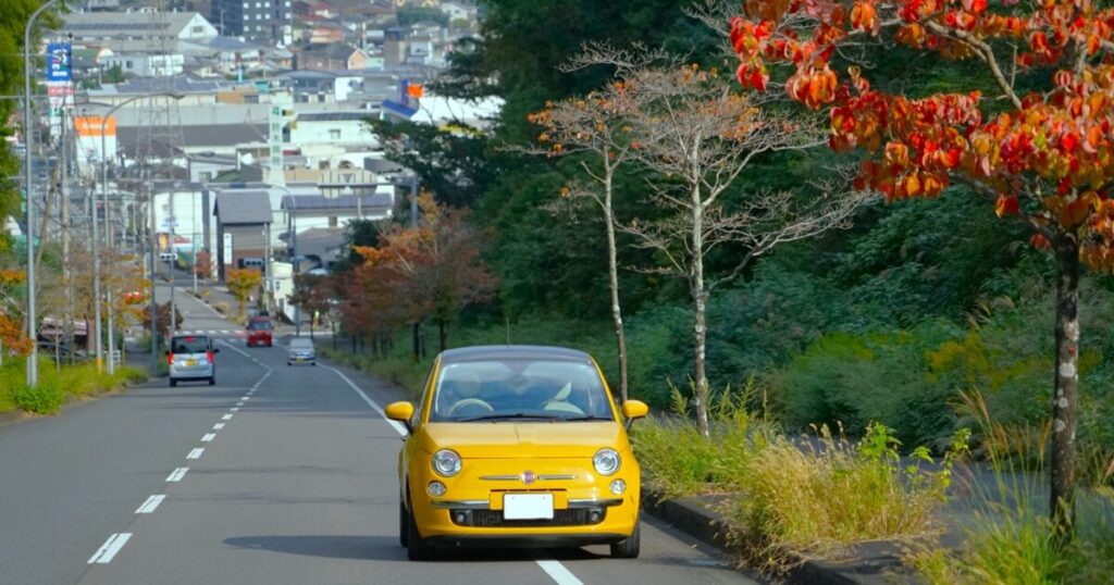 紅葉の並木道を走る黄色いコンパクトカー