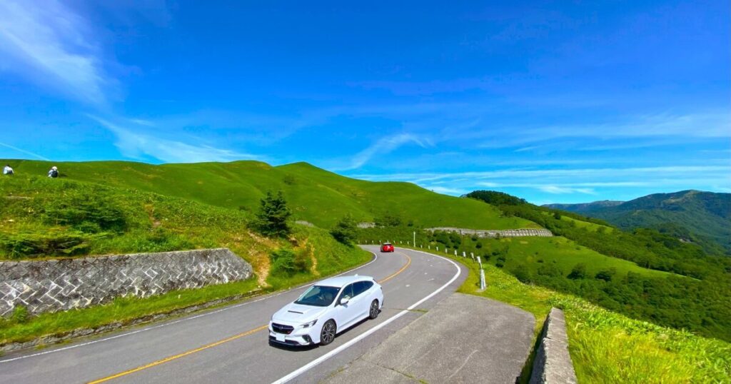 青空と緑の丘に囲まれたワインディングロードを走る白い車