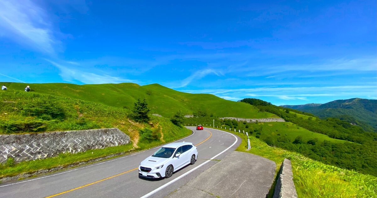 青空と緑の丘に囲まれたワインディングロードを走る白い車